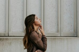 A young woman in a brown sweater stands next to a white wall, her hand under her chin and eyes closed, representing calmness and self-compassion.