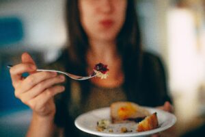 A blurred woman in the background holds up a white plate with a forkful of food, inviting a focus on mindful eating practices.